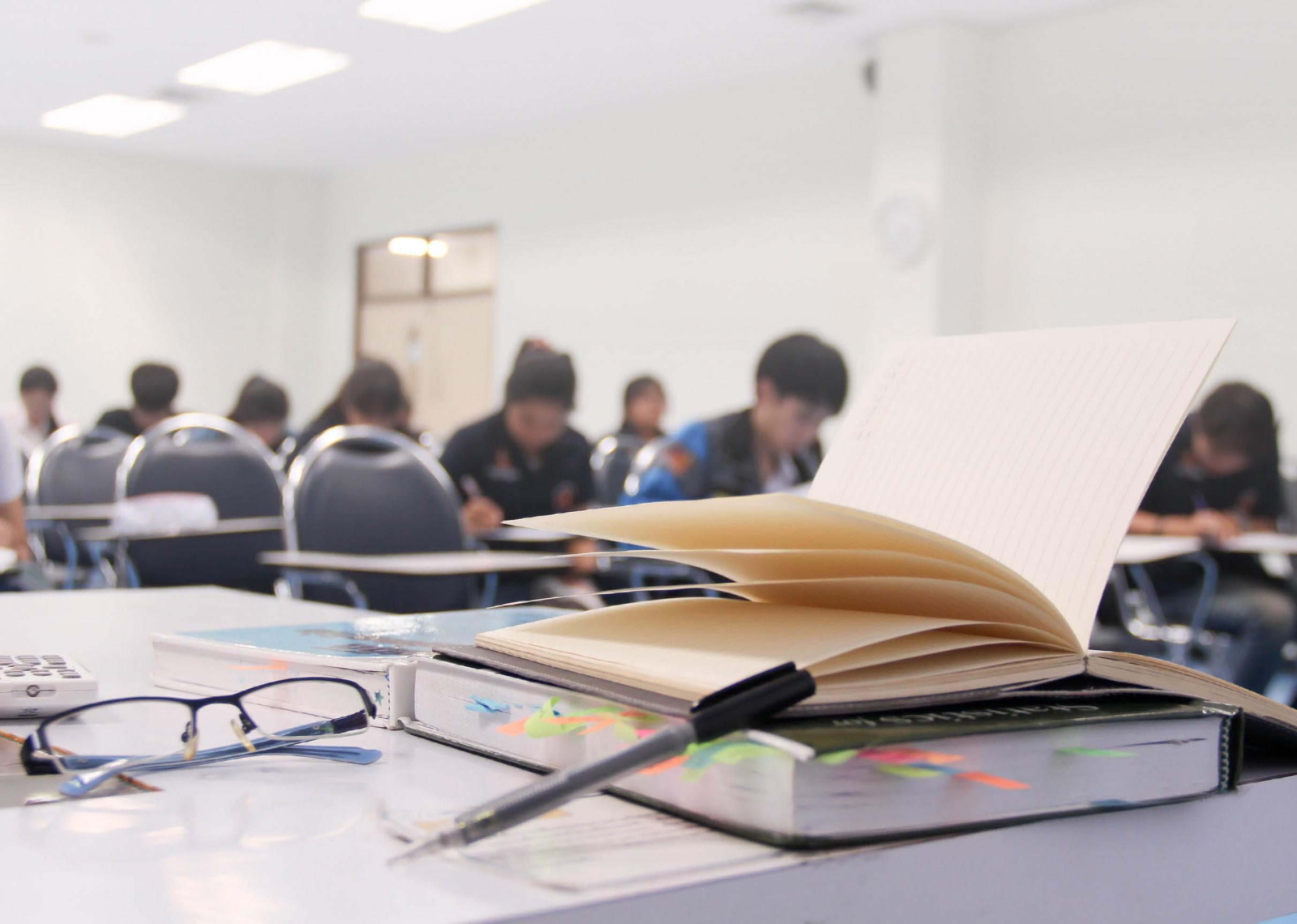 A notebook, pens, books and glasses on table and students in background