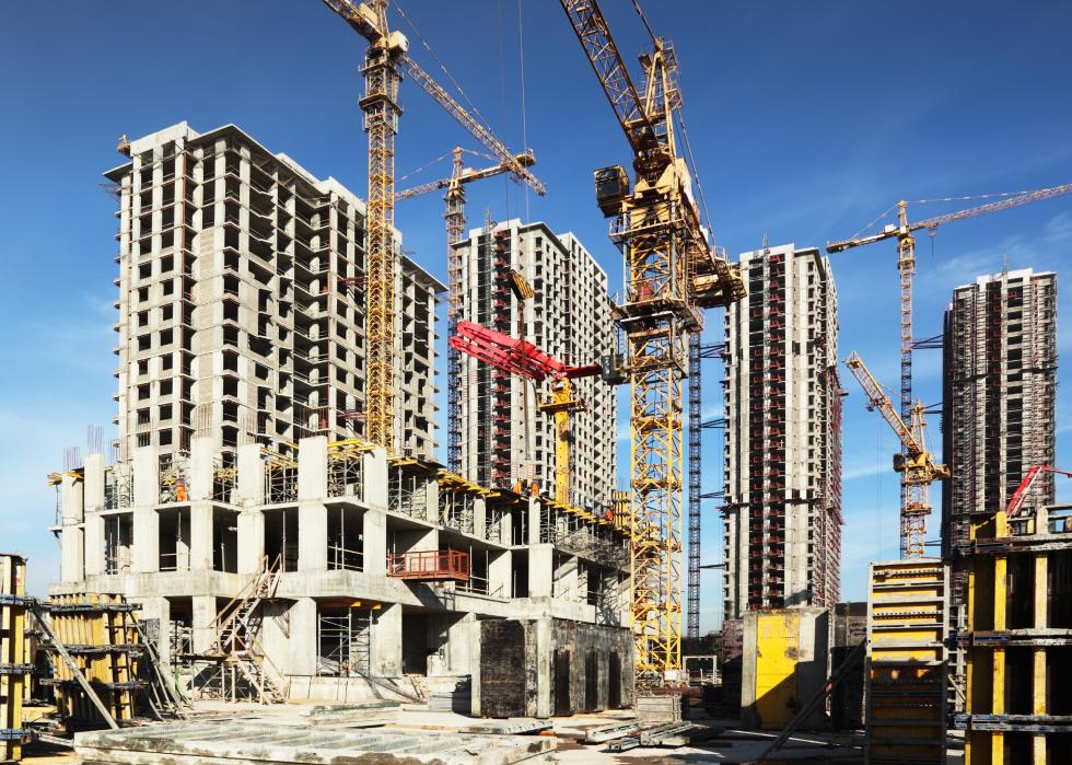 Several tall buildings under construction and cranes under a blue sky.