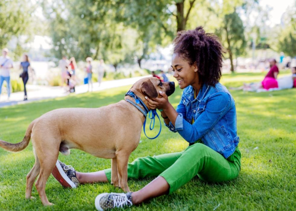 A smiling girl sitting on the grass, lovingly holding a medium-sized dog's head in her hands.