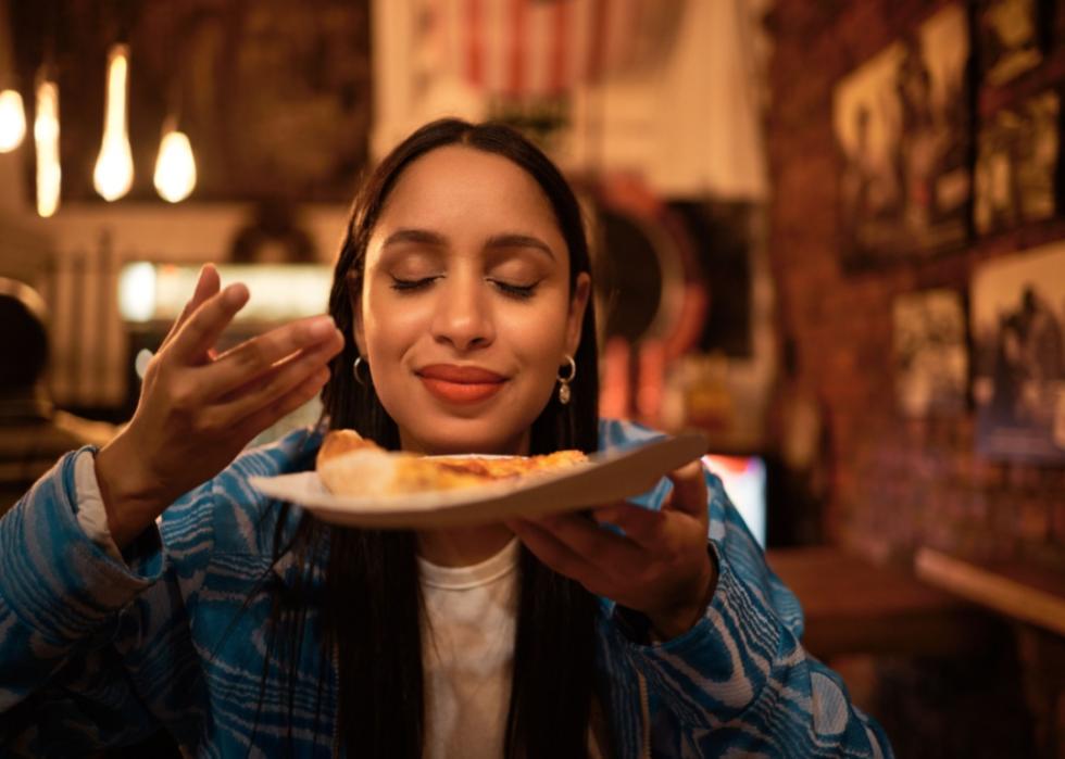 A person smelling the plate of food she is holding up to her face in a dimly lit restaurant.