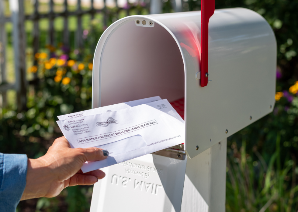 A hand placing a letter requesting a mail-in ballot in a white U.S. Mail mailbox