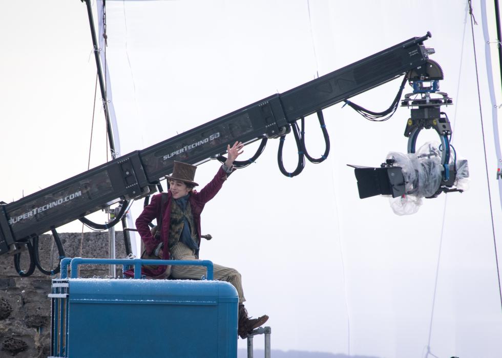 Timothee Chalamet dressed as Willy Wonka on the set of Wonka, sitting on a blue box next a large camera suspended in the air by a crane.