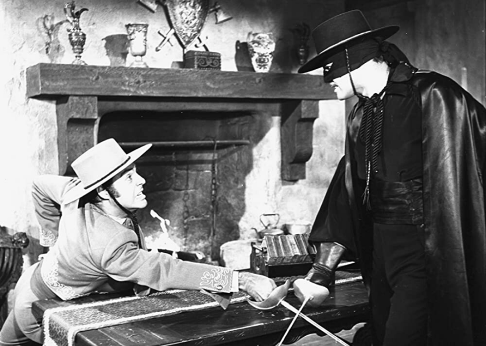 A man in a cape and mask sword fighting another man.