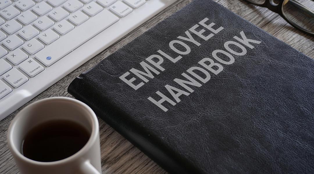An employee handbook and a cup of coffee in front a work computer keyboard.