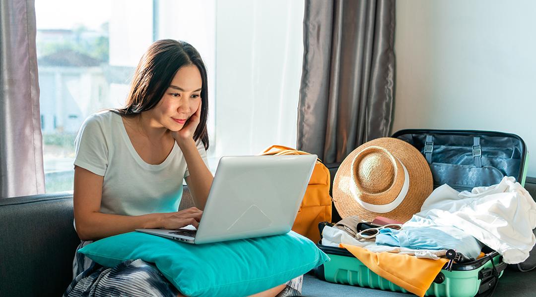 Young woman using a laptop beside an unpacked suitcase preparing for a trip.