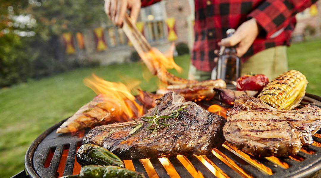 Assorted meat like sausages and steak with vegetables being grilled on a BBQ grill outdoors.
