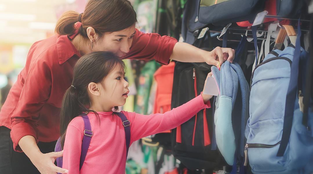 A mother helps her young daughter to pick a new bag for school at a store.