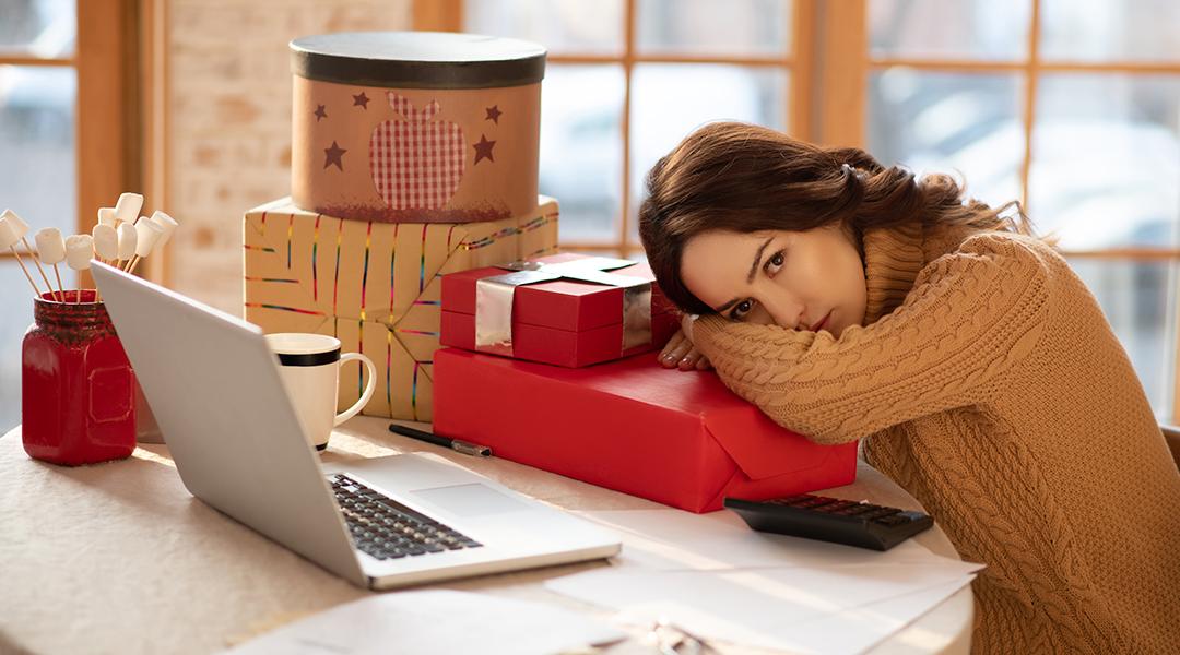 A young woman resting her head on a Christmas gift box alongside other presents in front of her laptop.