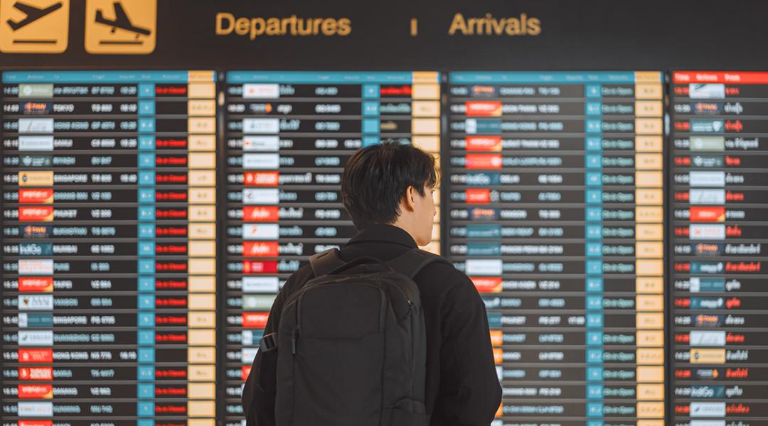 Rear view of a young man with a backpack looking at an airport's flights dashboard.