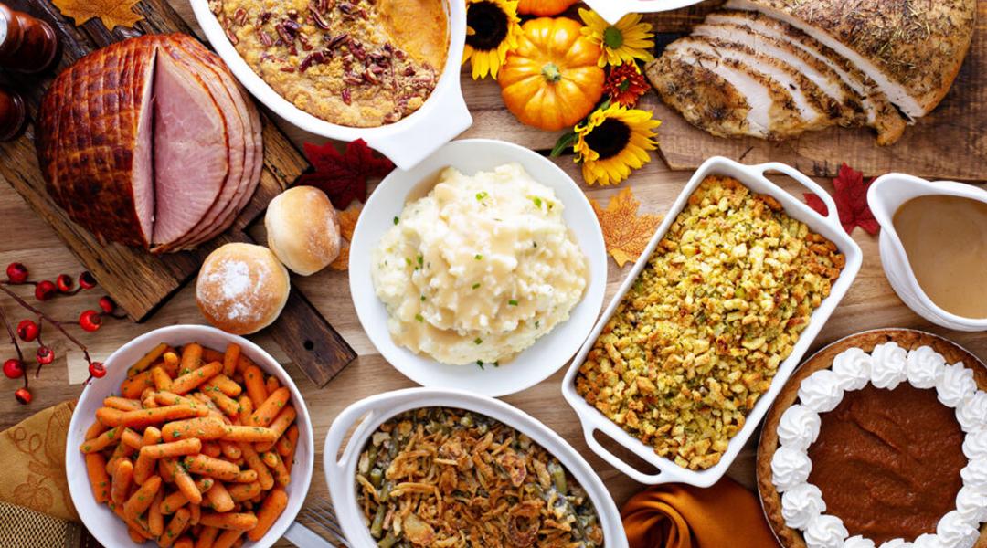 A table full of traditional holiday and Thanksgiving dishes such as mashed potato, ham, turkey, stuffing, and pumpkin pie.