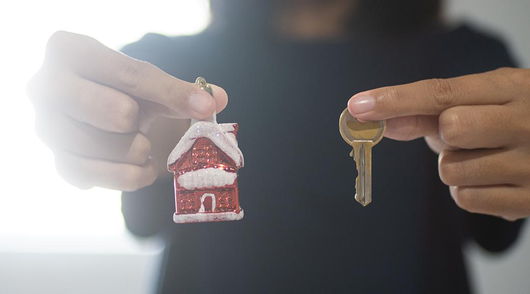 A woman holding a small figure of a Christmas home with her right hand and a house key with her left.