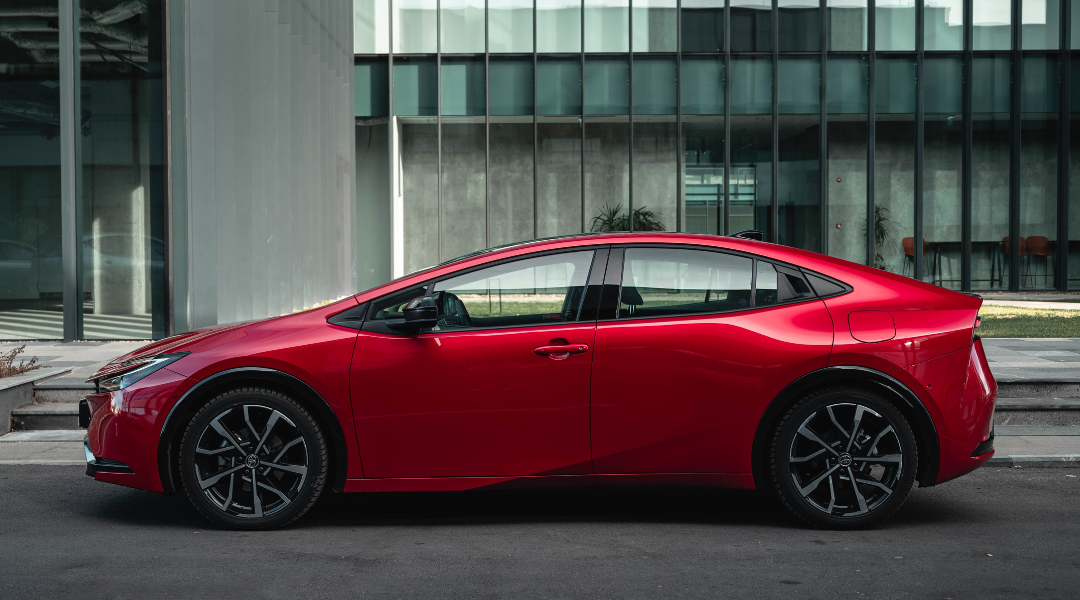A red 2024 Toyota Prius Prime parked in front of a building on a city street.
