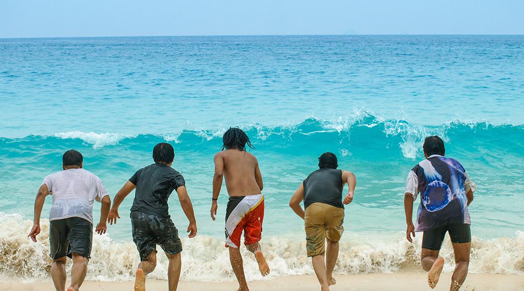A group of five friends lined up to run towards a sea wave from the beach.