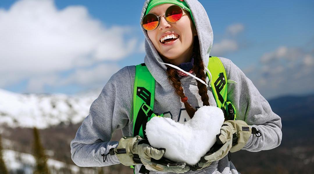A young woman in snowboarding gear holding a heart shape made of snow.