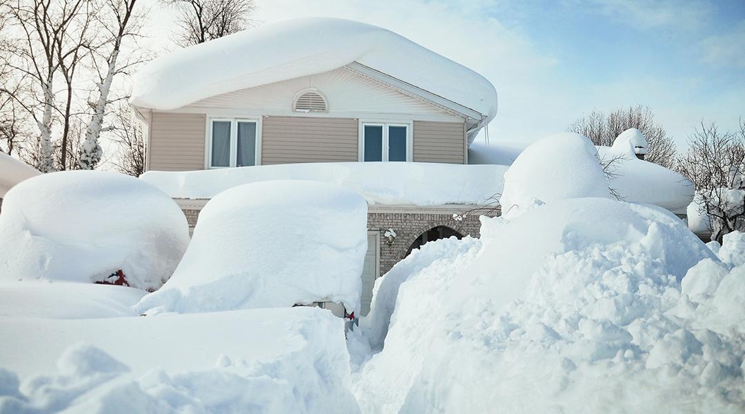 A house and its cars covered in deep white snow after a blizzard.