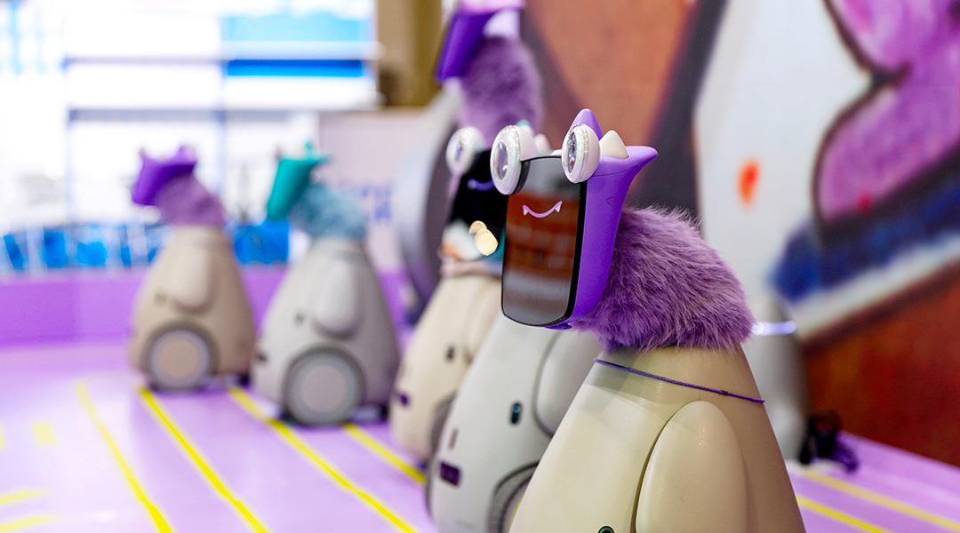 A row of 5 "robot pets" with playful googly eyes affixed on tablet faces and bright-colored faux fur necks on display at a convention. 