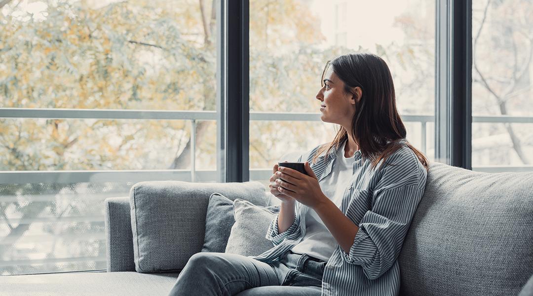 Young woman sitting on a sofa and having a cup of coffee while looking out the window at home.